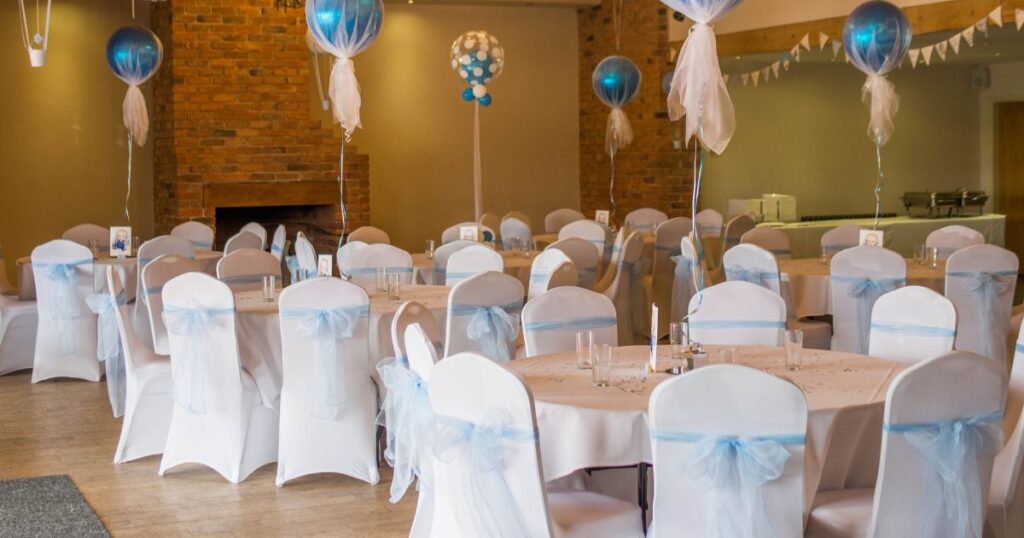 Elegant event setup featuring round tables with white and blue decor, chair covers, and balloon decorations in a banquet hall