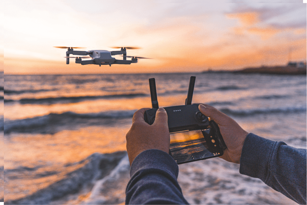 A person flying a drone over the ocean at sunset, capturing the scenic view with a remote controller. The drone hovers in the air, reflecting the warm hues of the sky and sea.